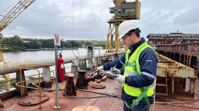 Profesional con casco, chaleco y arnés registra un relevamiento en una planilla sobre la cubierta de una embarcación en un astillero